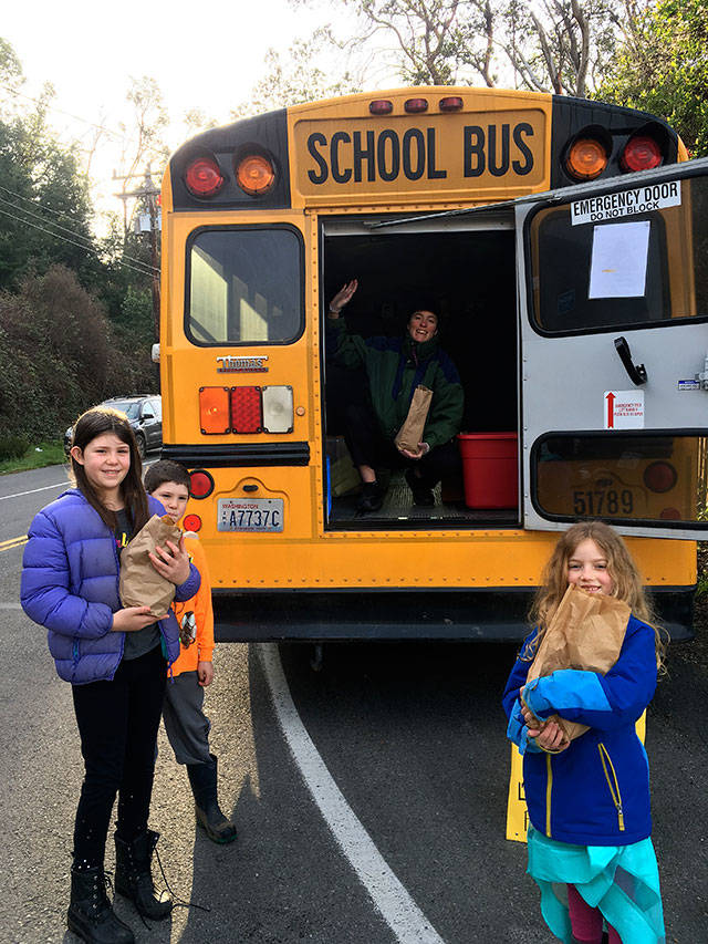 Vashon Island School District’s “grab and go” meal program includes pick-up at Vashon High School and about 15 spots around the island, with meals transported by school buses. Above, Holly Johnson, a district food service staff member, hands out food. She is joined by Liana Mann and Henry and Eleanor Casey at a Tahlequah bus stop (Courtesy Photo).