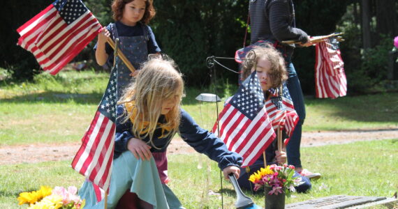 Tom Hughes Photo
Members of Vashon’s Cub Scout Pack 275, Luna Cekosh (kneeling, left), Hazel Ecevedo (standing), and Wilfred Gogarten (right) place American flags on the graves of veterans at Vashon Cemetery.