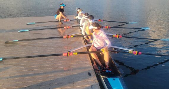 Ben Steele photo
Rose Ely, Gwen Tomlinson, Ren Colvos, Emily Rock, and Alekos Dalinis sit with rows at the ready. Coach Delany Steele crouches on the pier nearby.