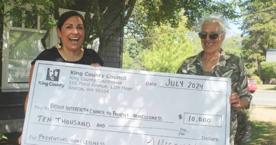 King County Councilmember Teresa Mosqueda (left) holds a big novelty check with the Vashon Interfaith Council to End Homelessness' Hilary Emmer, outside the Chamber of Commerce, on July 31. (Alex Bruell photo)