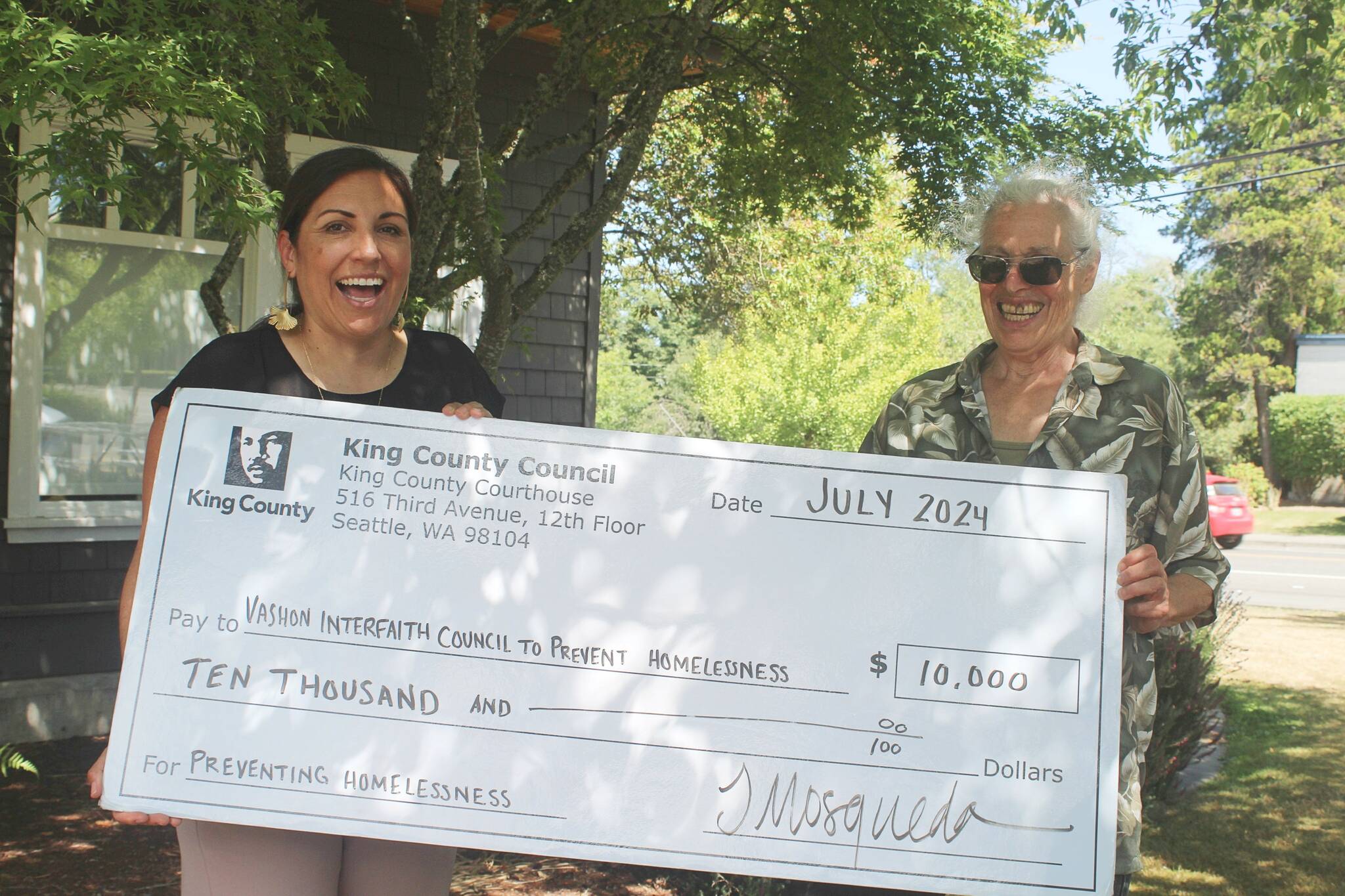 King County Councilmember Teresa Mosqueda (left) holds a big novelty check with the Vashon Interfaith Council to End Homelessness’ Hilary Emmer, outside the Chamber of Commerce, on July 31. (Alex Bruell photo)